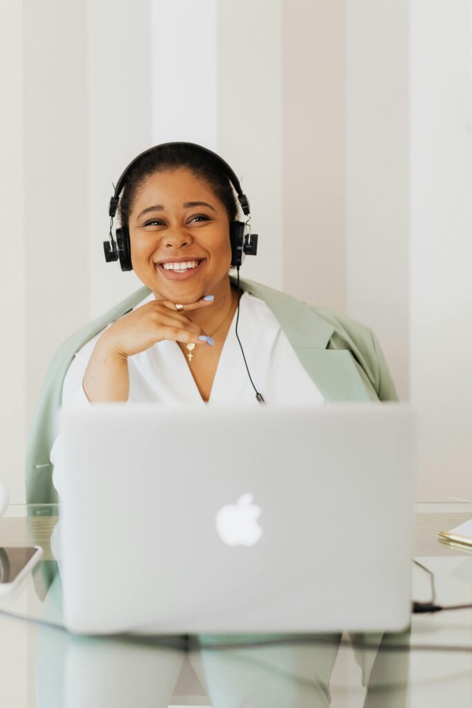 Black woman smiling while using a laptop, wearing headphones indoors.